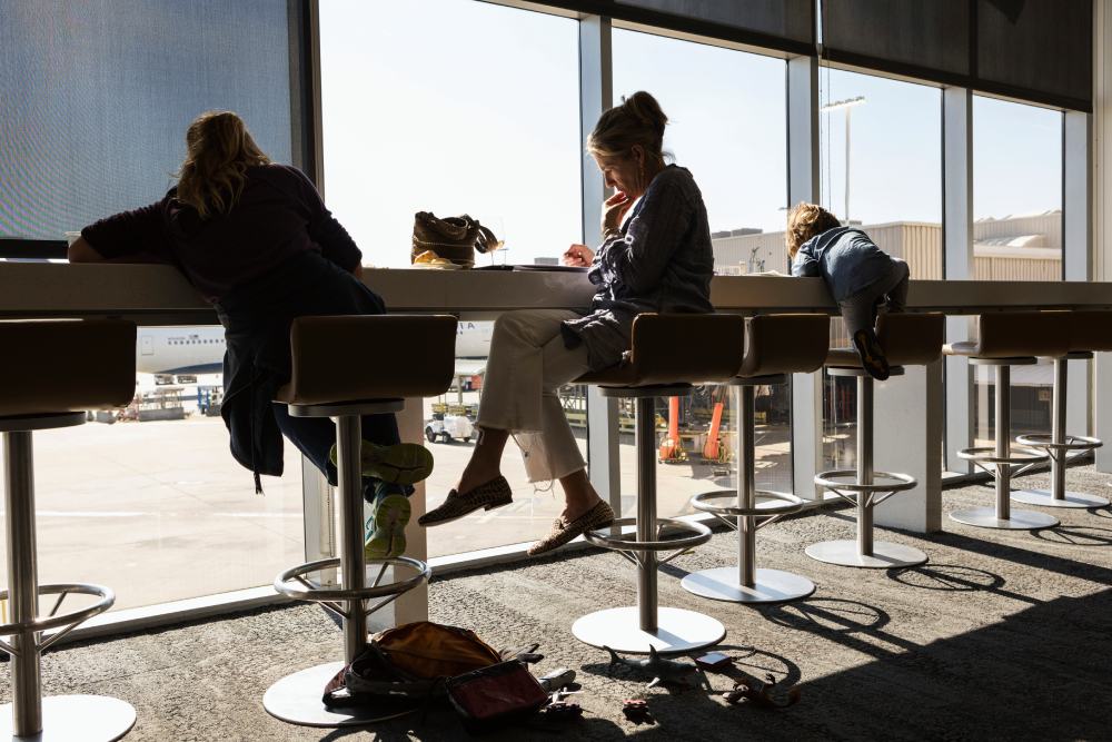 A mature woman and two children seated on high stools at an airport departure lounge iwth a view over the airport apron.
